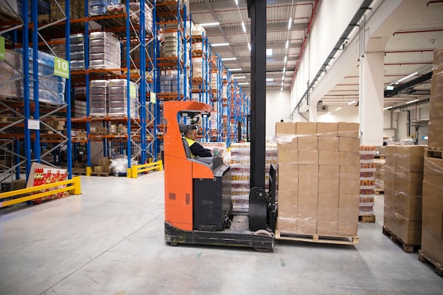 Free photo industrial worker in protective uniform operating forklift in big warehouse distribution center