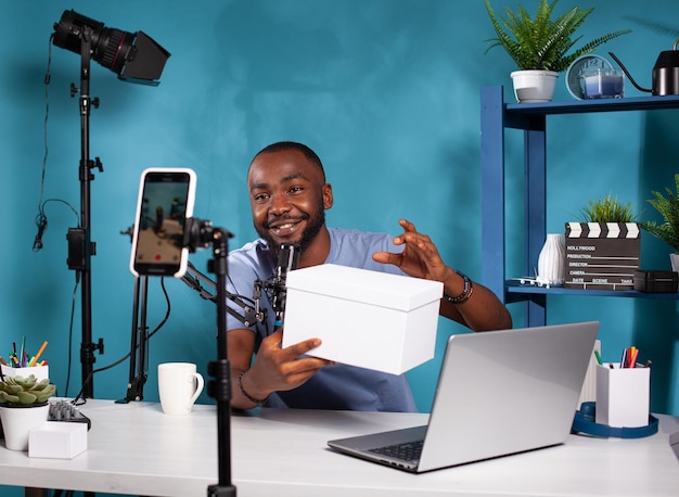 Free photo influencer presenting white mockup gift for fans looking at live video podcast setup in vlogging studio. smiling vlogger holding product box hosting online giveaway in front of recording smartphone.