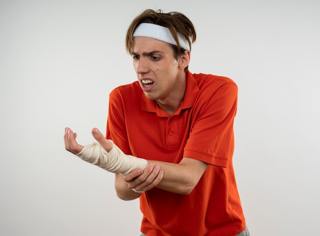 Free Photo injured young sporty guy wearing headband with wristband with wrist wrapped with bandage grabbed wrist isolated on white wall