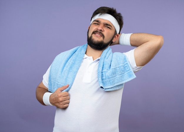 Injured young sporty man wearing headband and wristband with towel on shoulder grabbed aching neck isolated on green