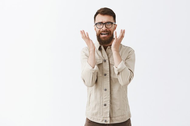 Insecure panicking bearded man in glasses posing against the white wall