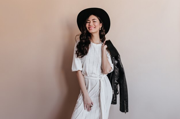 Inspired asian woman holding leather jacket. Studio shot of pretty chinese woman in dress and hat.