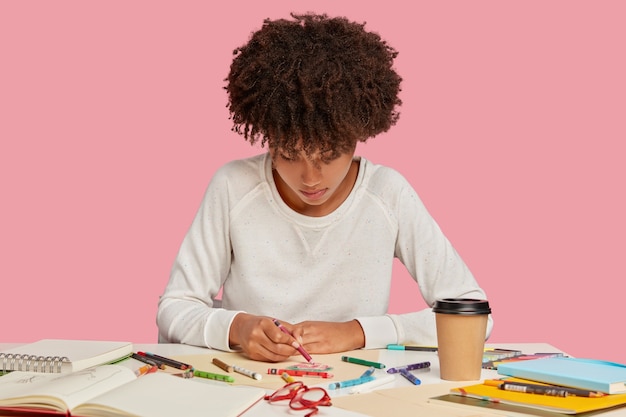 Free photo inspired black young woman enjoys drawing with crayons on blank sheet of paper, focused down, has mood for creativity, creats something original, sits at workplace alone against pink wall