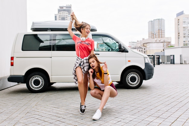 Free photo inspired brunette girl in yellow shirt sits with legs crossed beside white car while her friend playing with long hair