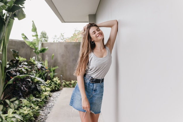 Free photo inspired female model with gorgeous hair posing outdoor near plants. portrait of shapely young woman in denim skirt standing beside white wall with romantic smile.