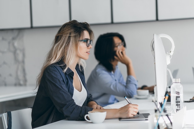 Inspired freelance web-designer using tablet and stylus, looking at screen while her friend talking on phone. Asian student holding smartphone and typing on keyboard, sitting beside blonde girl.