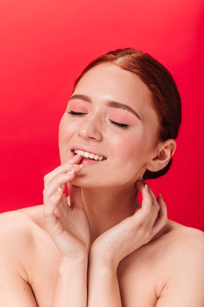 Free photo inspired ginger woman touching lips. studio shot of nude female model posing with closed eyes on red background.