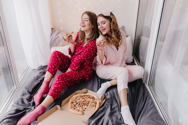 Inspired girl in pink socks eating pizza with best friend. Indoor photo of two sisters in pajamas enjoying italian food in bed.