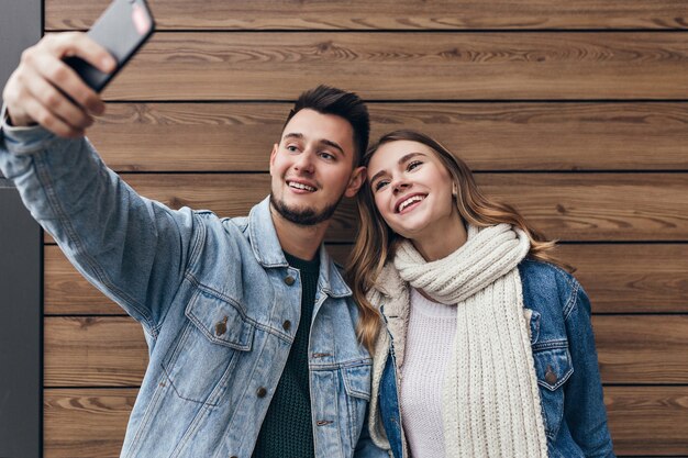 Inspired man with beard making selfie with his girlfriend. Gorgeous young woman with black scarf posing on wooden wall.