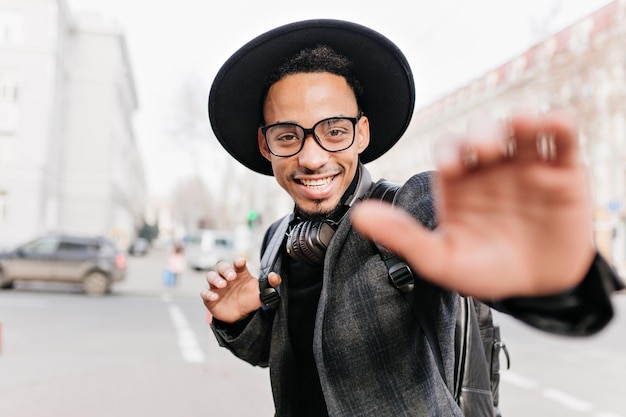 Inspired young african man with sincere smile posing on blur city background in morning. Handsome mulatto guy spending autumn day outdoor.