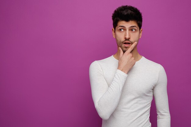 Inspired young handsome man keeping hand on chin looking at side on purple background with copy space