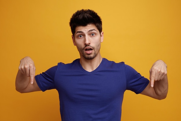 Inspired young handsome man pointing fingers down looking at camera on yellow background