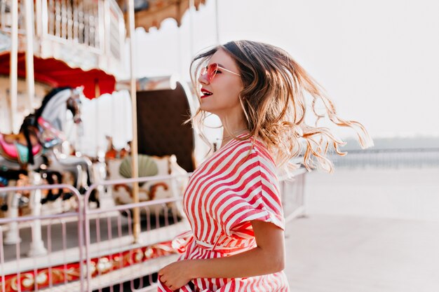 Inspired young woman dancing near carousel. Cheerful caucasian girl in striped dress expressing happiness in amusement park.