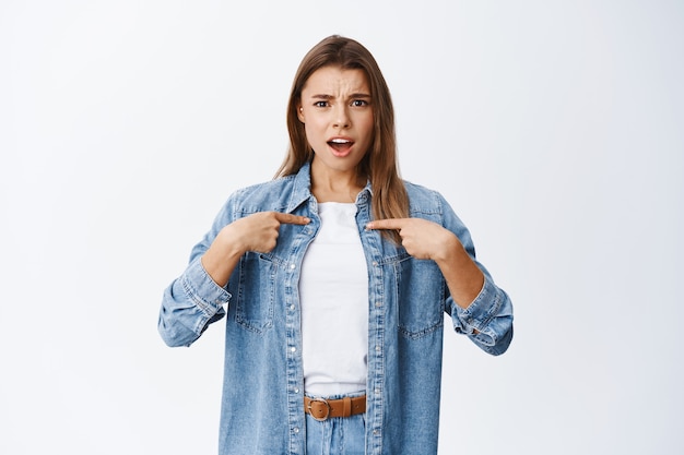 Insulted blond girl being offended, pointing at herself and frowning displeased, being accused, standing against white wall