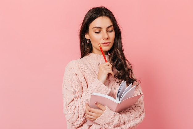 Intelligent woman in pink sweater thinks about sequel of her book. Brunette looks in her diary on pink background.