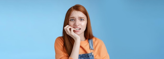 Free photo intense worried scared unsure young redhead panicking silly girl frowning look upset anxious biting fingernails emotional frightened fired standing blue background emotional terrified