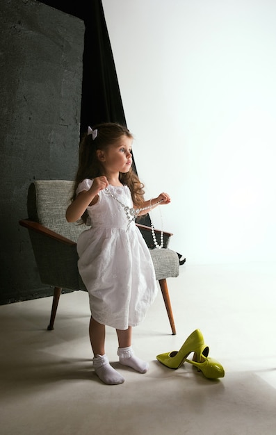 Free photo interaction of children with adult world. cute girl wearing mum's oversize shoes and dress for being older like she is. little female model trying on clothes at home. childhood, style, dream concept.