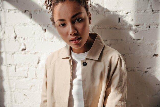 Interested african american woman in trench coat looking at camera Front view of curious black girl standing on bricked background