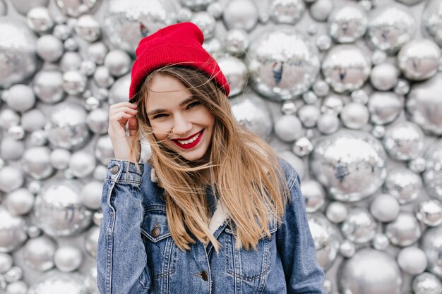 Interested beautiful girl in denim jacket touching her long hair. Smiling european female model in hat posing on shiny wall.