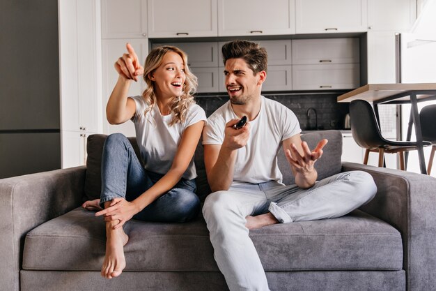 Interested blonde lady watch TV. Indoor portrait of smiling couple sitting on cozy couch.