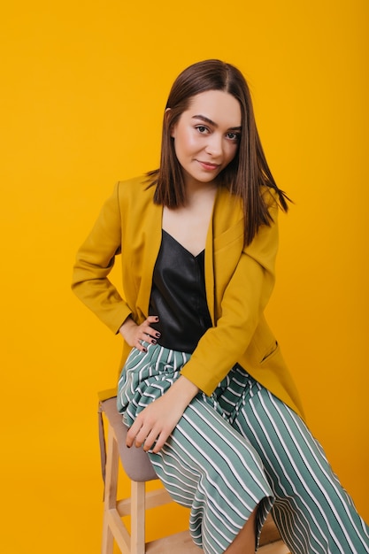 Interested brunette girl in bright jacket sitting on chair. lovely brown-haired woman in striped pants posing with pleasure.