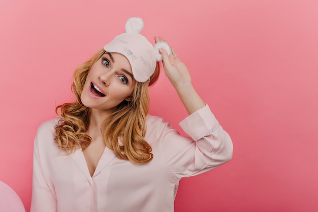 Free photo interested curly woman with big eyes laughing. dreamy young female model in eyemask expressing surprised emotions on bright wall.