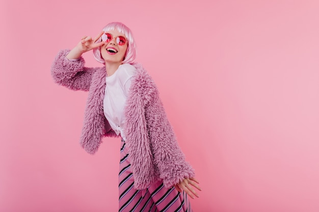 Free photo interested funny girl in periwig and sunglasses posing with peace sign. indoor shot of blissful caucasian woman wears fluffy jacket and pink peruke