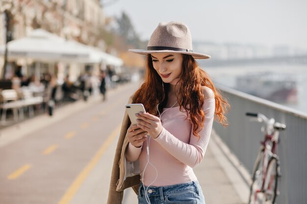 Interested ginger woman with phone in hands checking mail on city wall