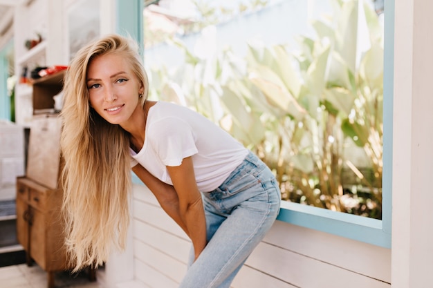 Interested lightly-tanned woman posing in sunny morning. Indoor photo of fashionable blonde girl standing near window.