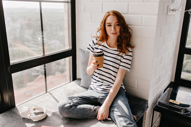 Free Photo interested white lady with cup of coffee looking. joyful red-haired girl sitting near window.