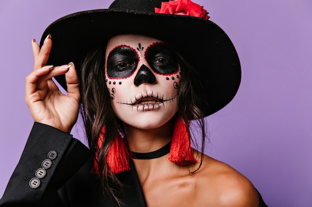 Free photo interested woman with scary face painting. halloween portrait of brunette latin girl in big black hat.