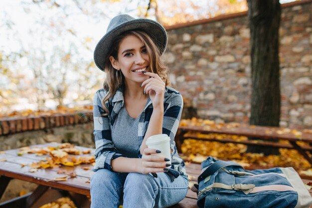 Interested young lady with cup of coffee posing in park in october day