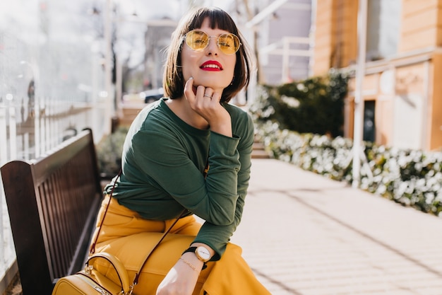 Free photo interested young woman in yellow pants sitting on the street. outdoor photo of winsome brunette girl posing on bench.