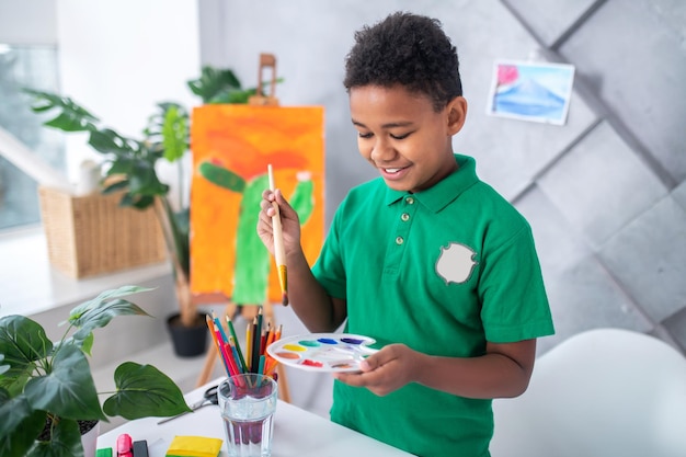 Interesting activity. Smiling dark-skinned boy of primary school age in green tshirt looking holding paintbrush over palette with paints standing near table with pencils and glass of water