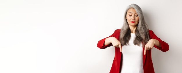 Free photo intrigued asian elegant woman looking and pointing fingers down showing logo standing in red blazer