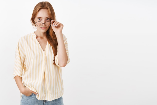 Free photo intrigued smart woman with ginger hair and freckles taking off glasses and looking under rim with curious expression smirking hearing interesting news