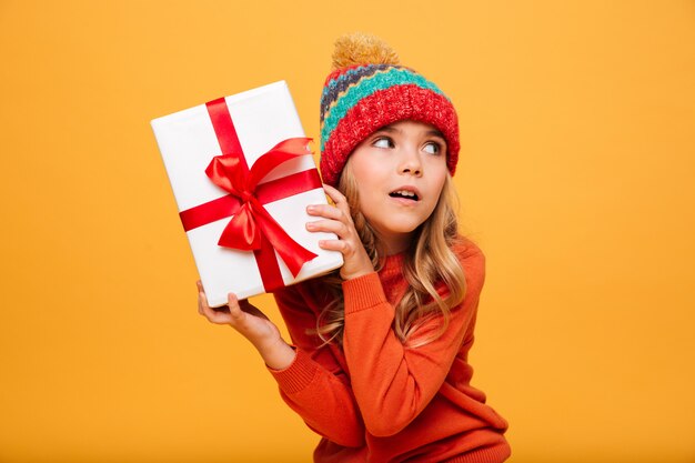 Intrigued Young girl in sweater and hat holding gift box and looking away over orange