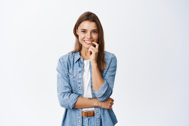 Intrigued young woman touching lip and smiling seeing something interesting looking thoughtful and pleased standing against white background