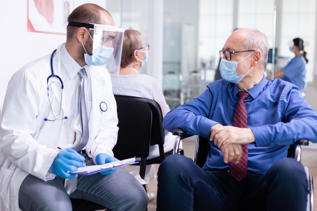 Free photo invalid old man with face mask against infection with coronavirus in wheelchair discussing with doctor in hospital waiting area