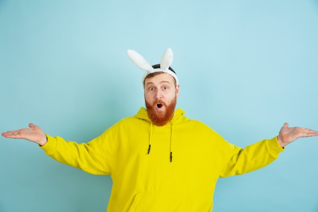 Free photo inviting, greeting. caucasian man as an easter bunny with bright casual clothes on blue studio background. happy easter greetings. concept of human emotions, facial expression, holidays. copyspace.