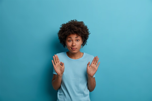 Do not involve me in this. Portrait of clueless dark skinned woman raises palms, doesnt want to get into trouble, doesnt feel guilty after doing something wrong, isolated on blue wall.