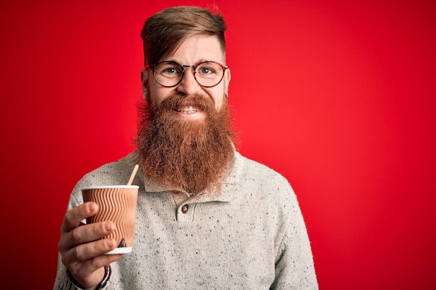 Irish redhead man with beard drinking a takeaway coffee on a paper cup over red background with a happy face standing and smiling with a confident smile showing teeth