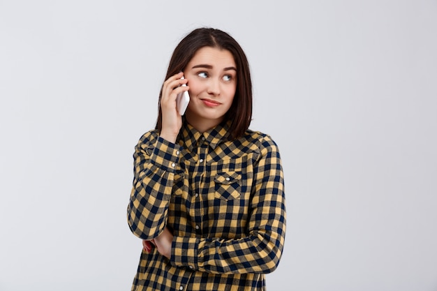 Ironic young beautiful girl dressed in plaid shirt speaking on phone, looking at side over white wall.