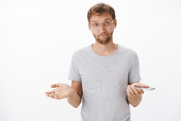 Free photo irritated handsome guy with brislte in glasses shrugging with spread hands in clueless gesture listening song in wireless earphones holding smartphone