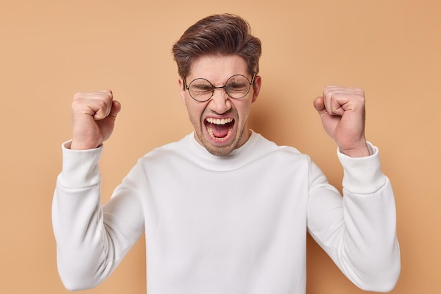 Free photo irritated outraged man expresses negative emotions keeps fists clenched screams loudly keeps mouth opened dressed in casual jumper isolated over beige background being in fury. i hate you all