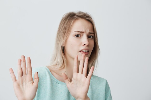 Irritated scared blonde young female with straight fair hair in blue sweater posing against wall, keeping hands in stop gesture, trying to defend herself, saying: Stop that. Body language.