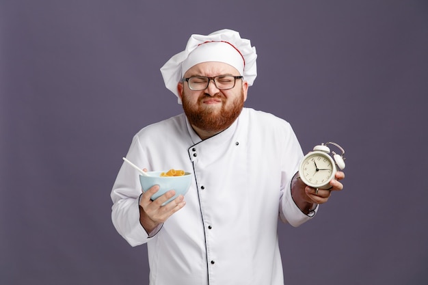 Irritated young chef wearing glasses uniform and cap holding alarm clock and bowl of macaroni pasta with spoon in it with eyes closed isolated on purple background