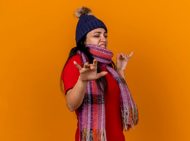 Irritated young ill woman wearing winter hat and scarf standing in profile view keeping hands in air with closed eyes isolated on orange wall