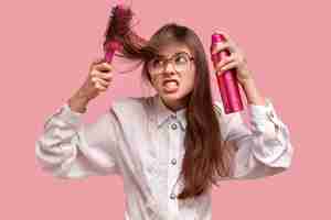 Free photo irritated young lady makes hairstyle with hairspray and hairbrush, has ratty hair