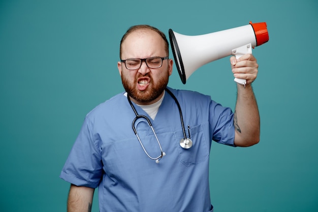 Free photo irritated young male nurse wearing glasses nurse scrub and stethoscope around his neck pointing speaker at head with closed eyes isolated on blue background